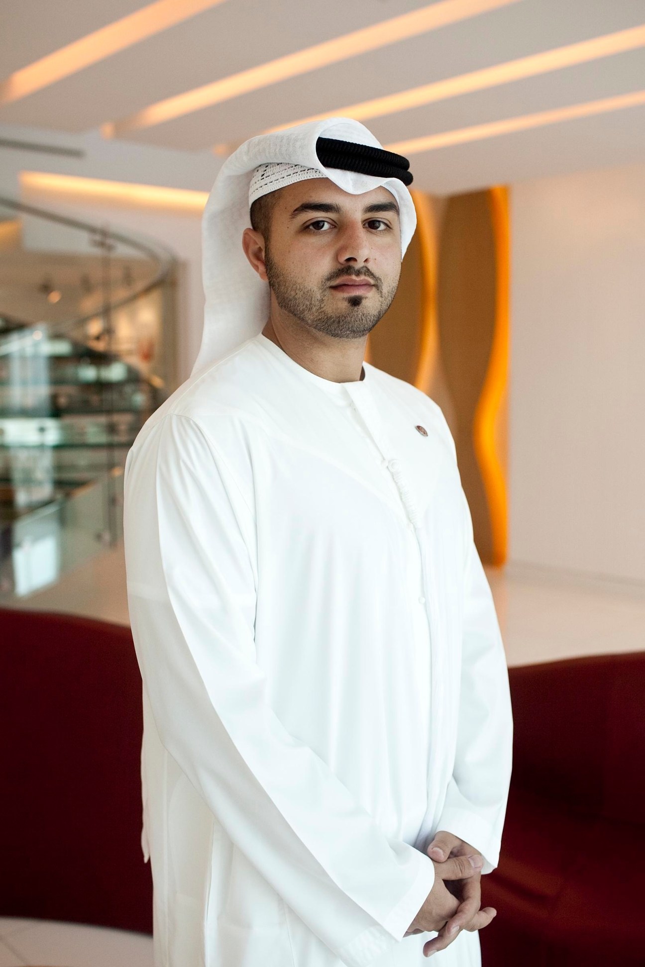 Man in white traditional Emirati dress and white ghutra standing in modern office with warm lighting.