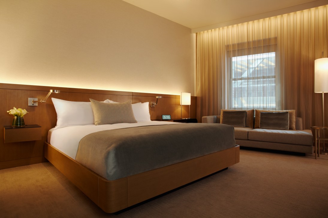 A modern hotel bedroom with warm wood tones, white bedding, and contemporary furnishings overlooking a cityscape.