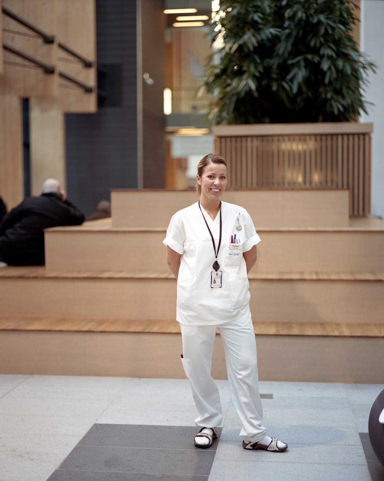 Nurse Kjersti Sykepleier stands smiling in the modern atrium of Akershus University Hospital, with wooden architectural details and plants visible behind her.