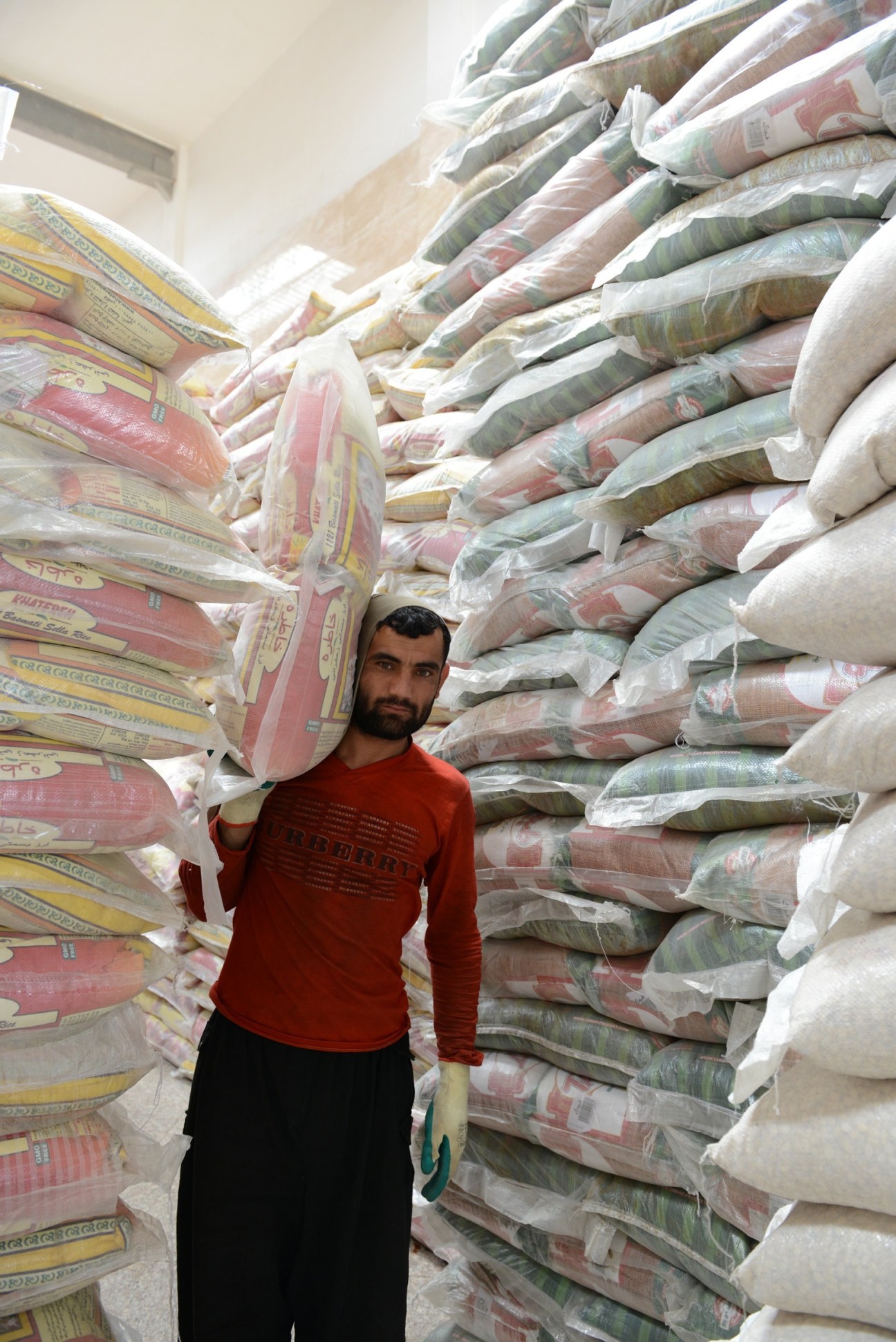 Man in red shirt standing between tall stacks of packaged carpets in a storage warehouse.