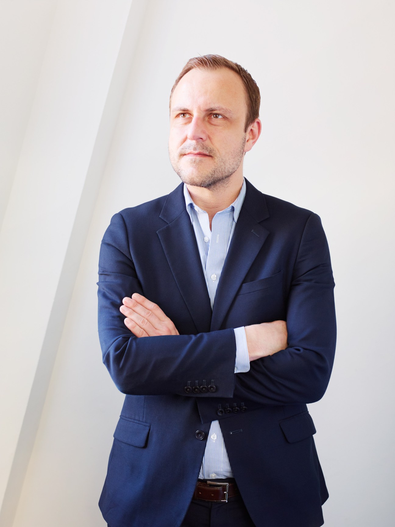 Portrait of Peter Neumann, a man in a dark blazer and light blue shirt with arms crossed against a white background.