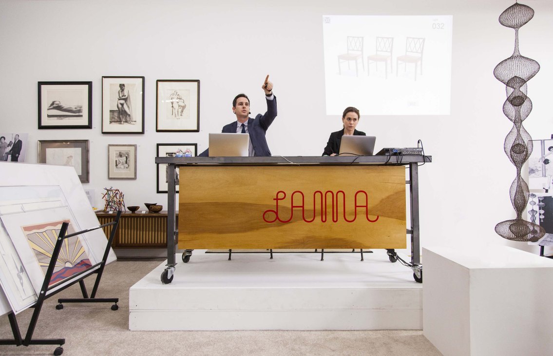 Two auctioneers at a wooden podium branded with 'LAMA' during a modern art and design auction in Los Angeles.