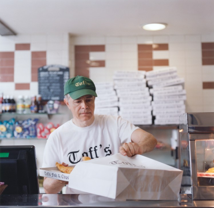 Staff member at Toff's fish restaurant wearing green cap and white shirt with Toff's logo, holding a fish and chips order.