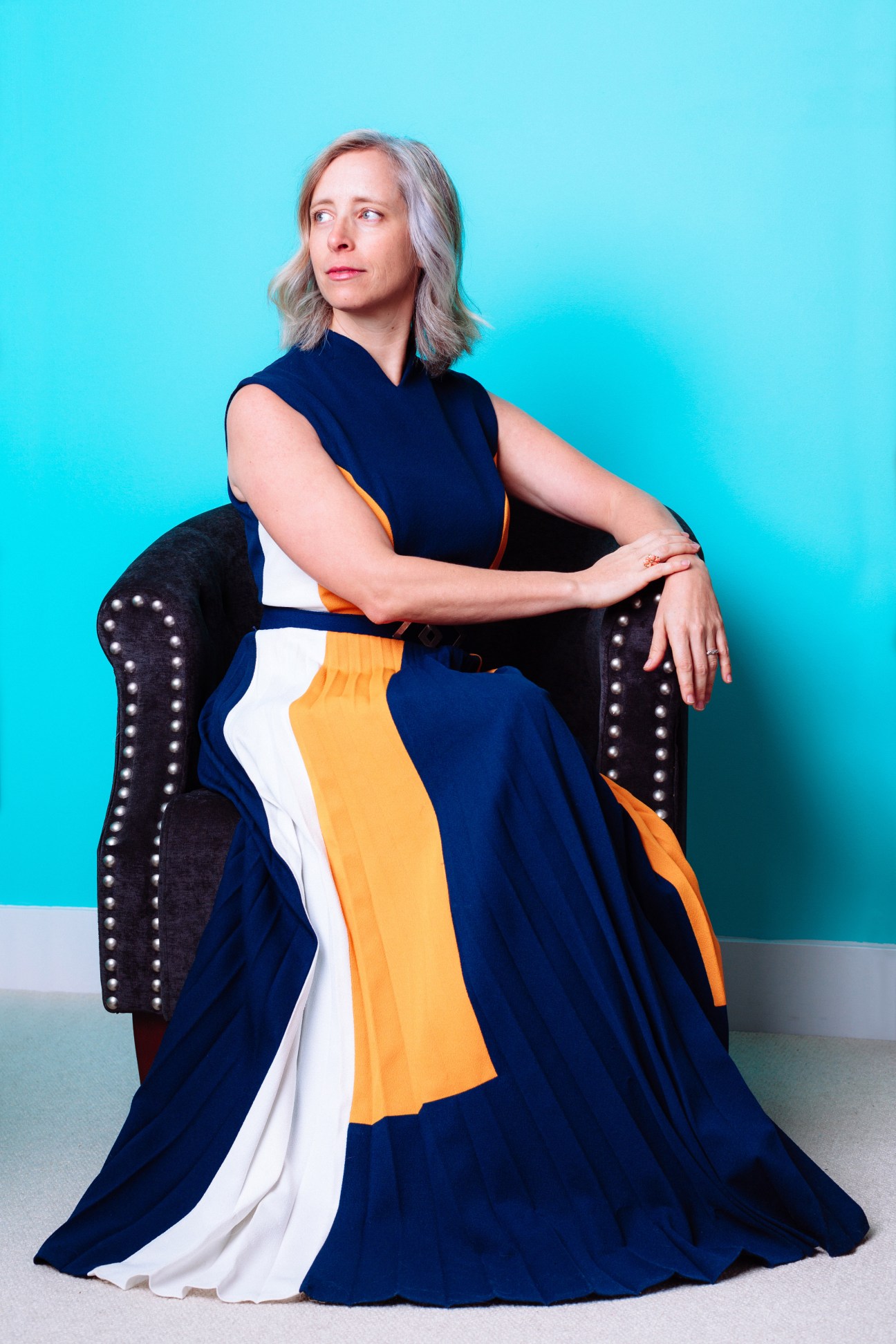 Woman with shoulder-length grey hair seated in a black studded armchair against a turquoise background, wearing a navy and orange striped dress.