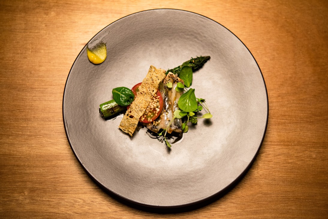 Grilled mackerel fillet with crispy skin, asparagus, tomato, and fresh herbs on a white plate.