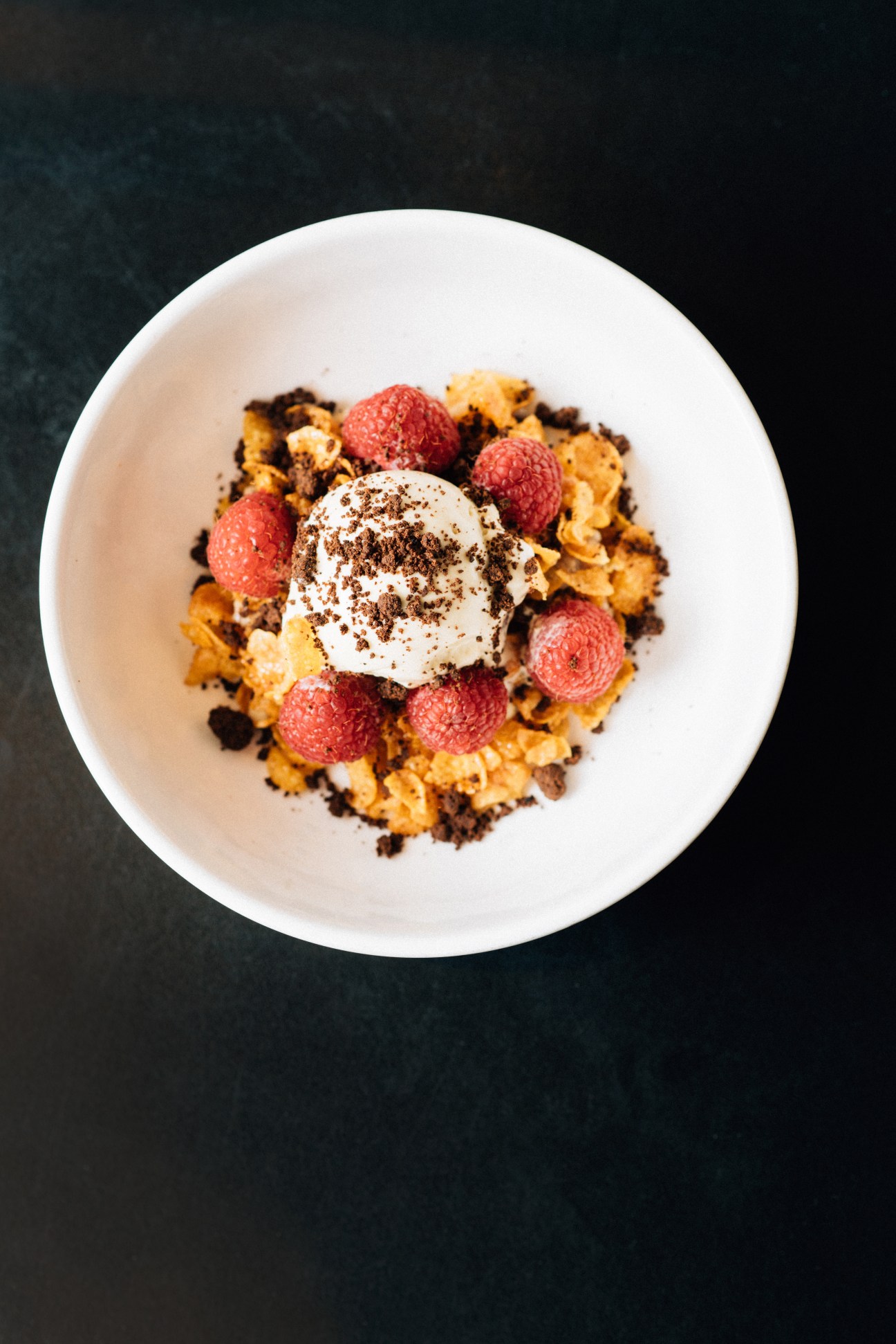 A white bowl containing oatmeal porridge topped with Rocky Road ice cream, fresh raspberries, toasted cornflakes, and coffee crumbs.