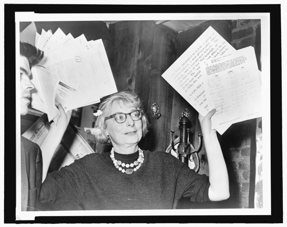 Black and white photograph of a woman with glasses and pearl necklace holding papers at a microphone.