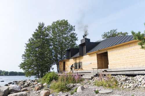 Modern sauna building with black roof and wooden exterior on Lonna island, Helsinki, with lake and trees in background.