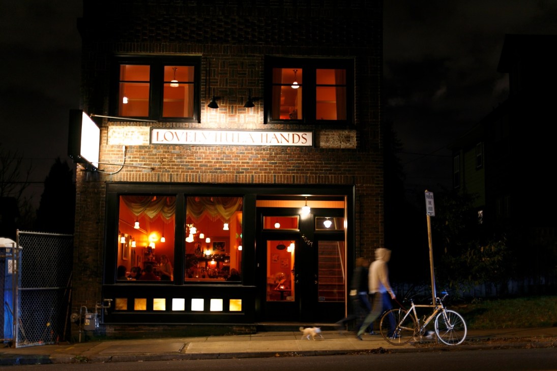 Lovely Hula Hands restaurant storefront at night with warm lighting and a cyclist passing by.