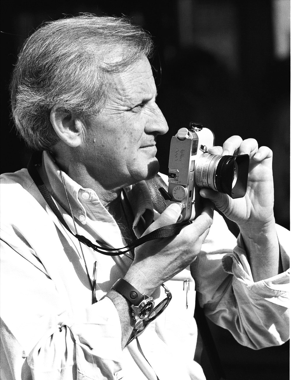 Man in profile holding a film camera with lanyard, wearing light-colored shirt against dark background.