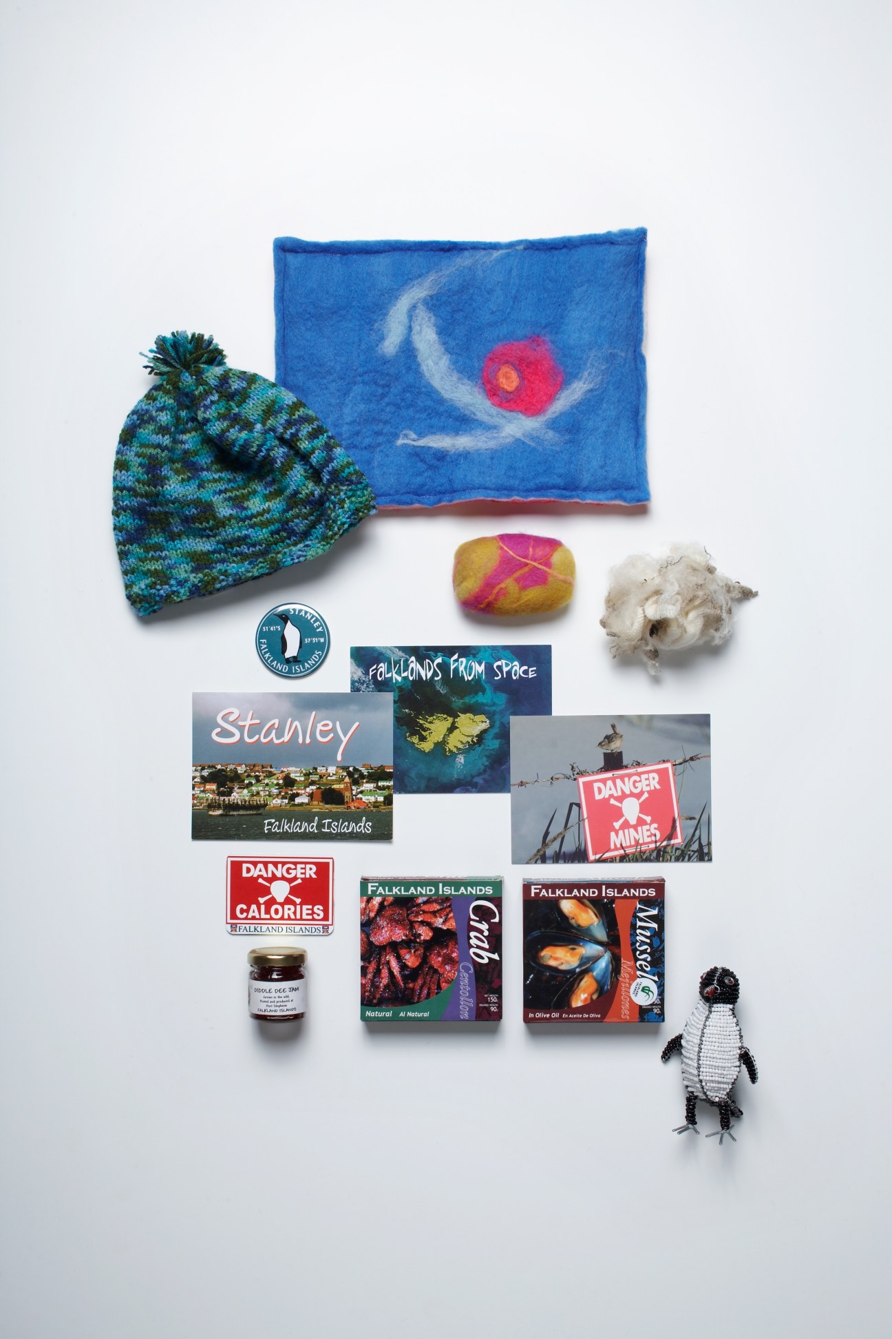Assortment of Falkland Islands products including wool hat, felt crafts, postcards, canned foods, jam, penguin ornament, and souvenirs arranged on white background.