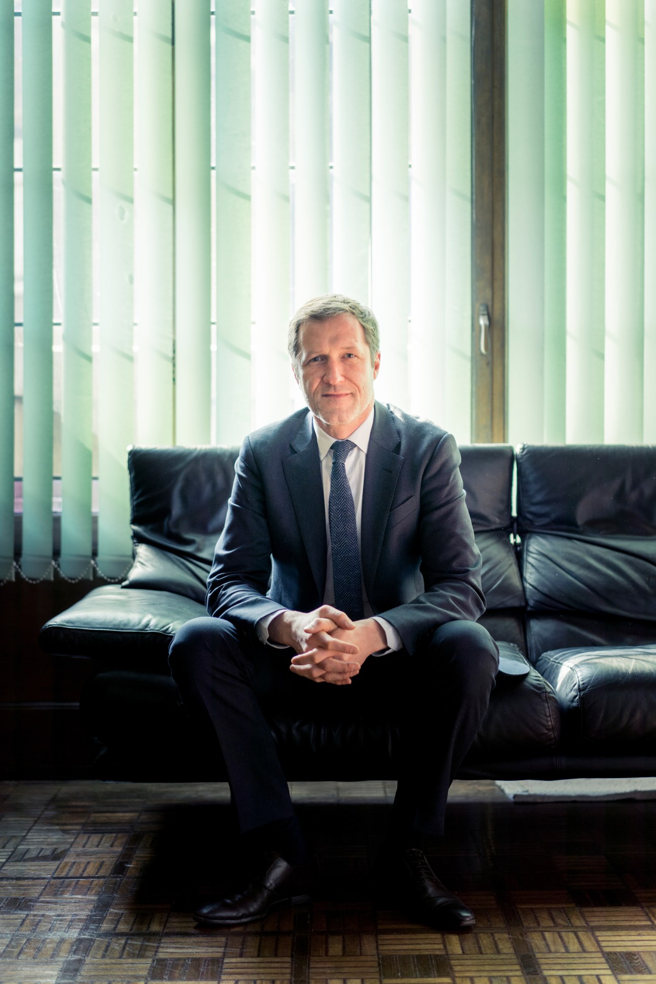 Paul Magnette, Mayor of Charleroi, seated on a black leather sofa against a green striped backdrop.