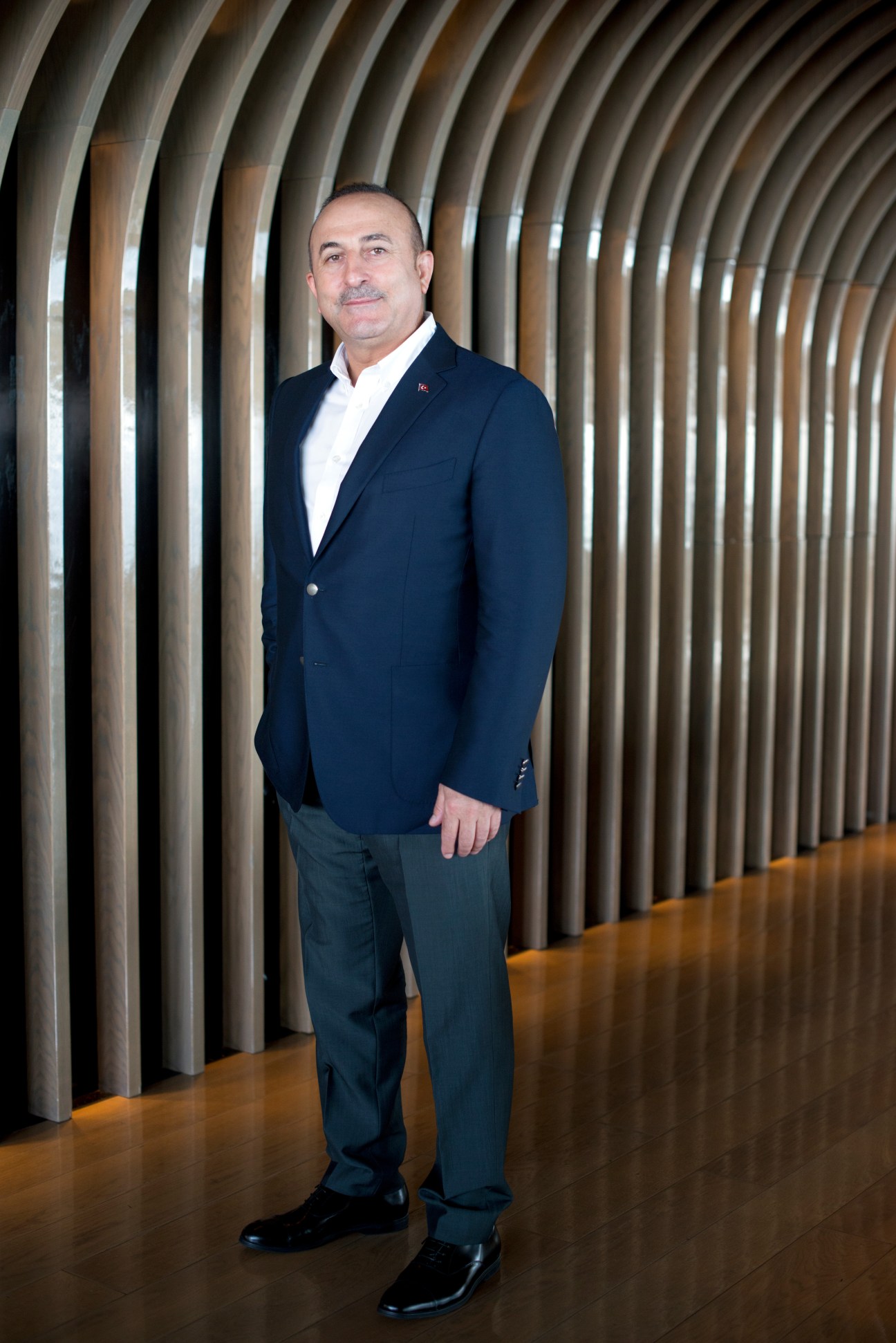 Mevlut Cavusoglu, Turkey's foreign minister, stands in a modern interior with curved architectural elements behind him.