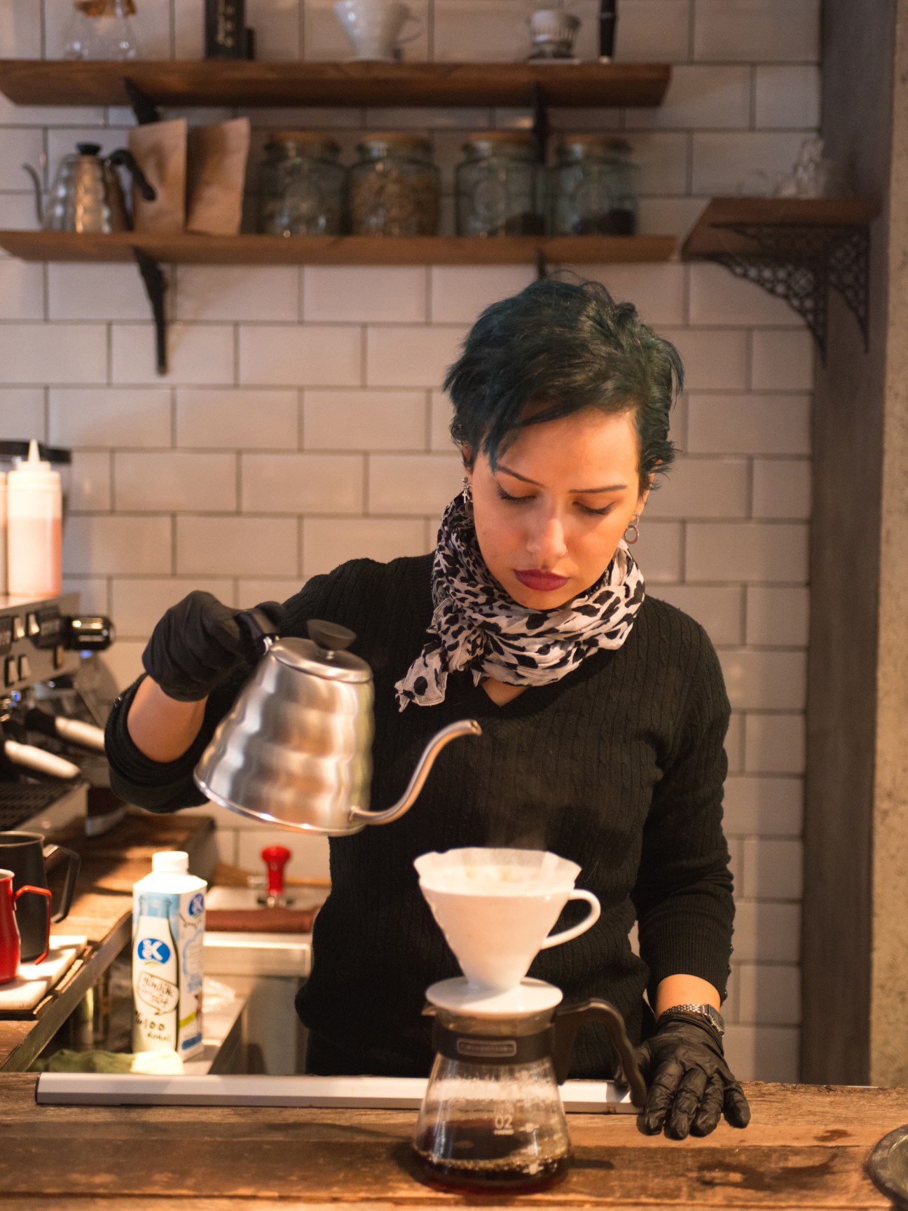 A barista pouring hot water from a metal kettle into a white pour-over coffee filter.