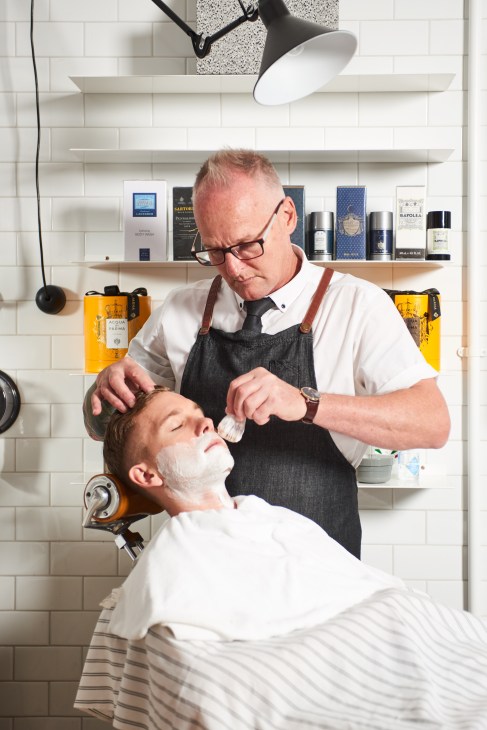 A barber applies shaving cream to a customer's face at Men's Biz barbershop with white subway tiles and product shelves visible behind.