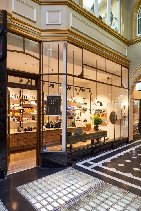 Men's Biz barbershop storefront in a Victorian arcade with glass windows and wooden interior displaying grooming products.