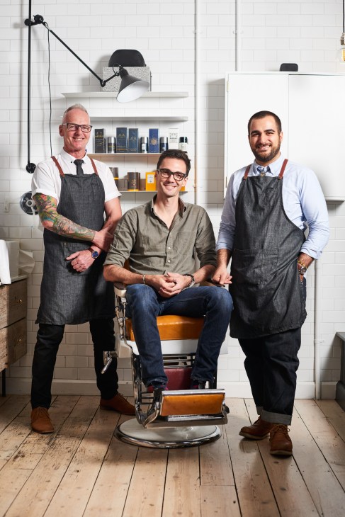 Three men posing in a barbershop with white subway tiles, shelves of grooming products, and a vintage barber chair.