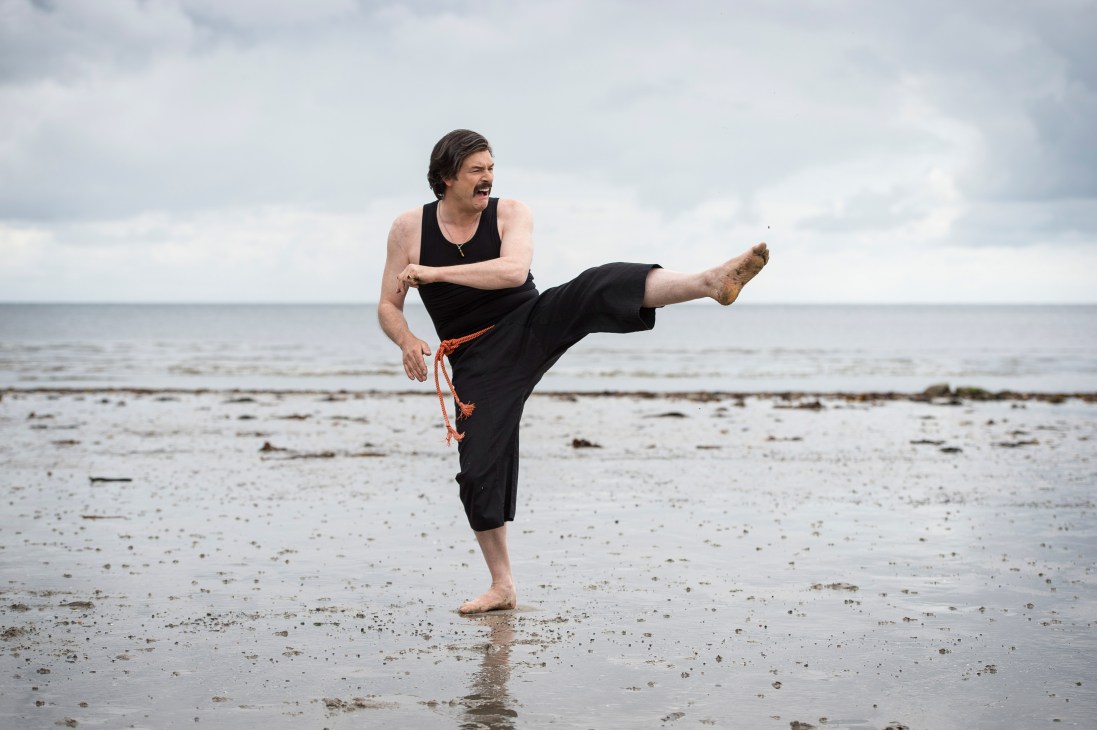Man in black tank top performing martial arts kick on beach with overcast sky.