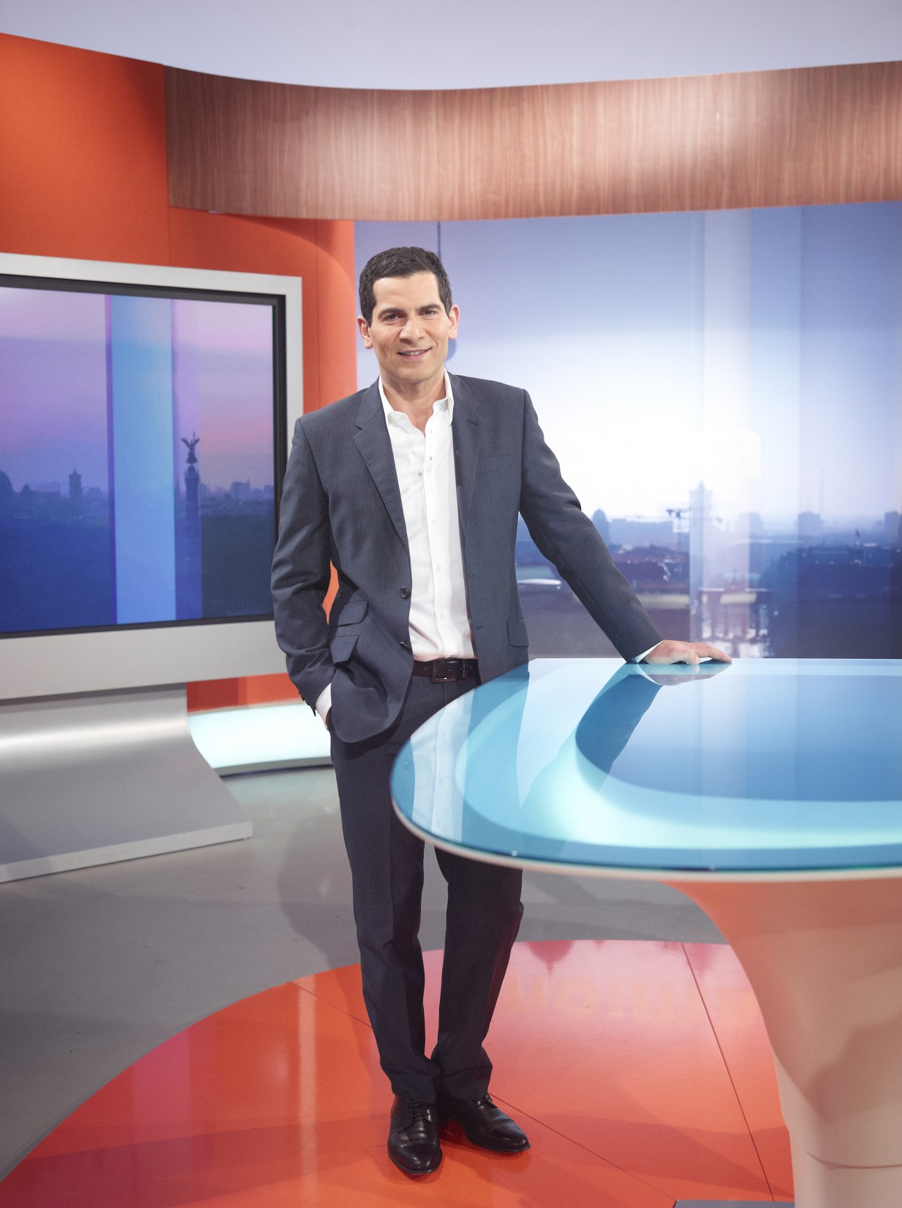 Mitri Sirin, host of Morgenmagazin, standing in a modern television studio set with orange and blue elements.