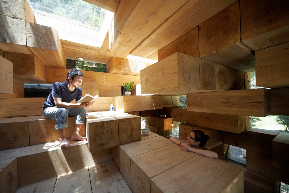 Two people in a multi-level wooden interior with stacked box-like rooms and skylights allowing natural light.