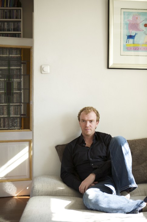 Man with reddish-blonde hair sits on a beige couch in a bright, minimalist interior with bookshelves and artwork.