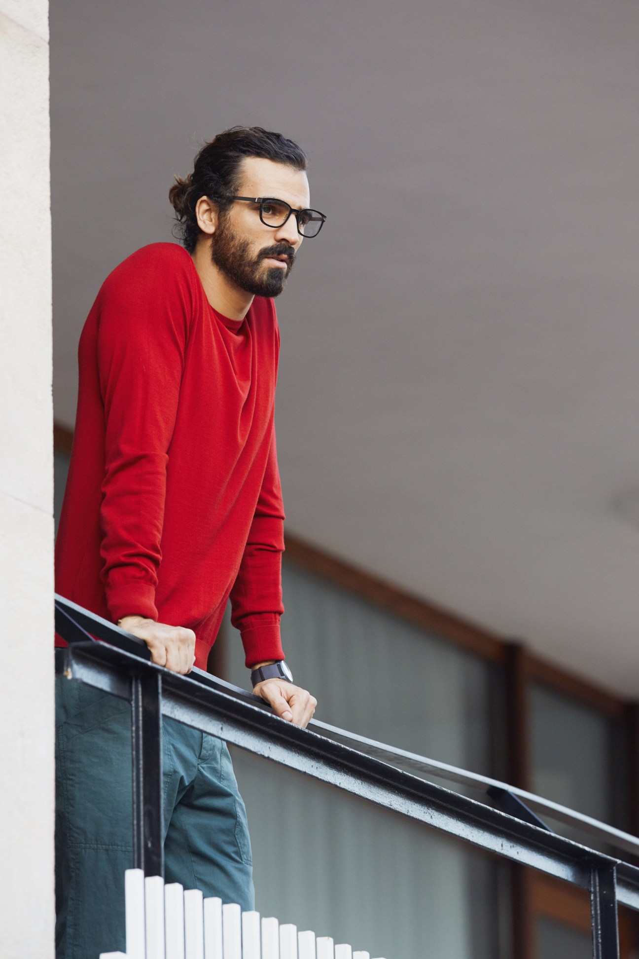 Man wearing red sweatshirt and dark trousers leaning against white wall with glasses