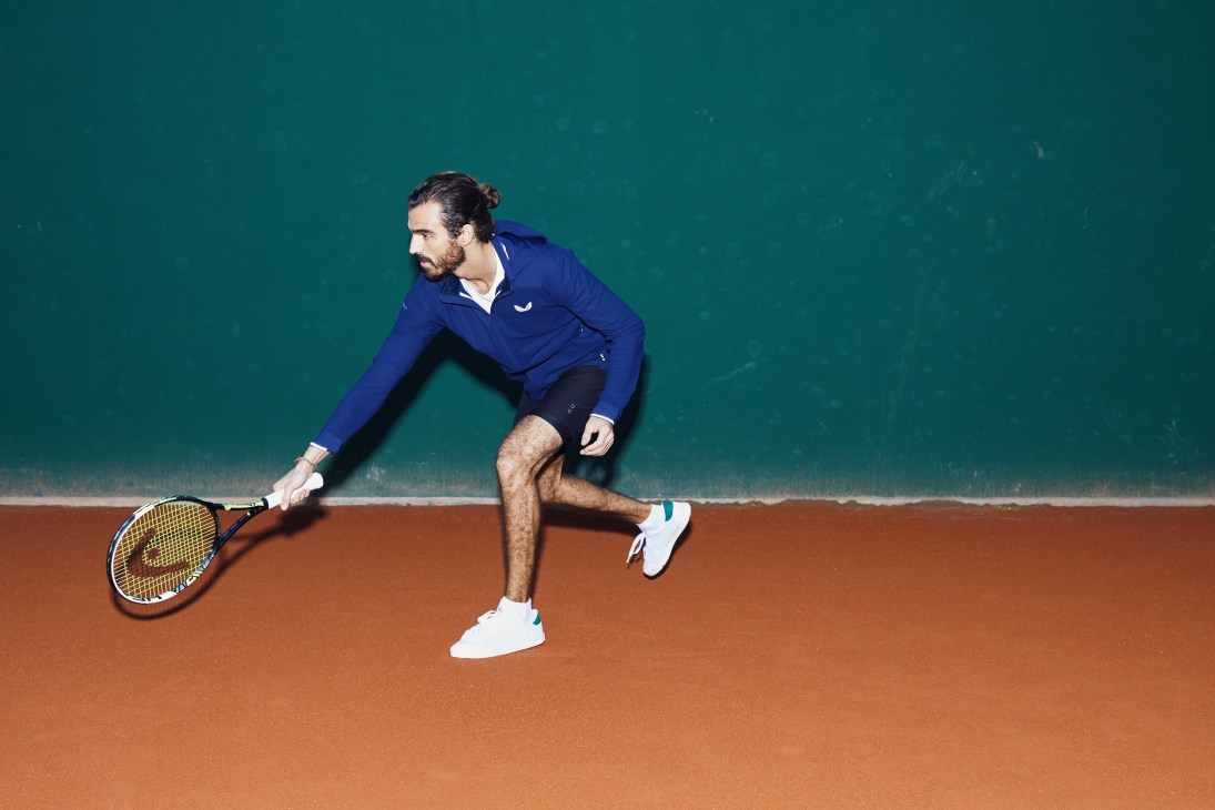 Man playing tennis on clay court wearing Castore jacket, Tracksmith t-shirt, and Ron Dorff shorts.