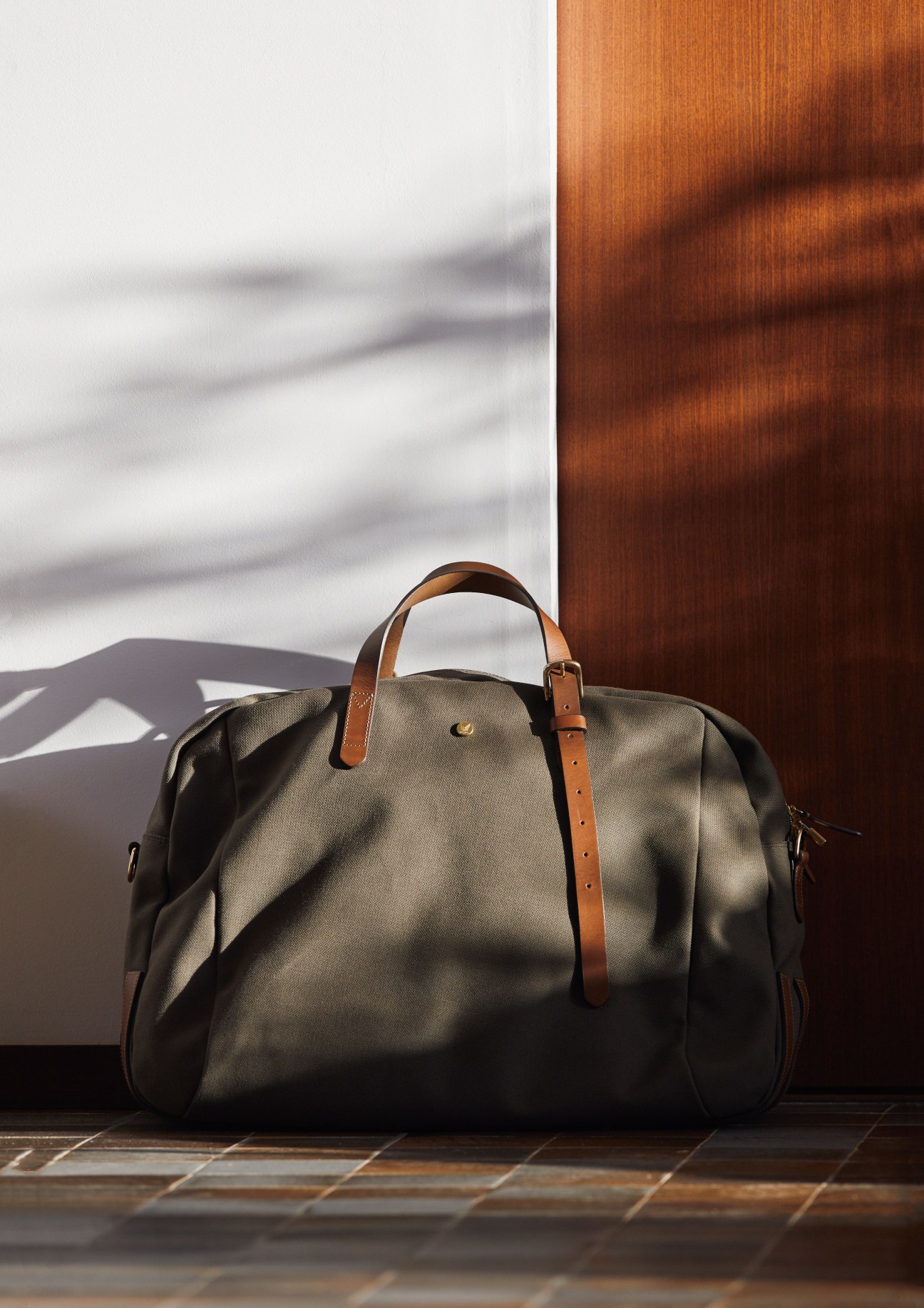 Khaki canvas travel bag with cognac leather handles and brass hardware against wooden wall.