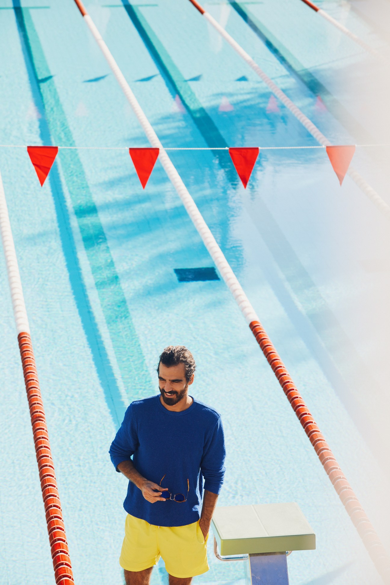 Man in blue long-sleeve shirt and yellow swim shorts standing in an empty swimming pool with lane dividers.