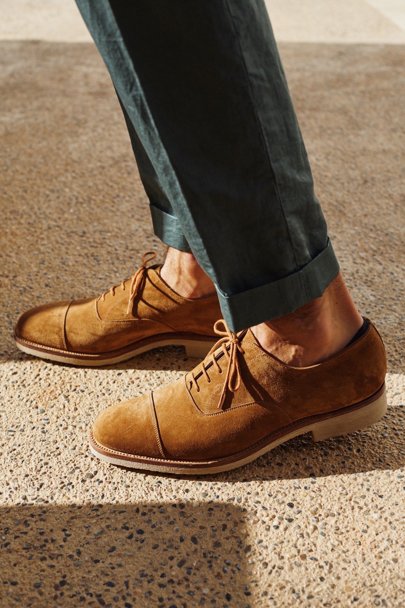 Close-up of tan suede oxford shoes and rolled dark jeans on concrete ground.