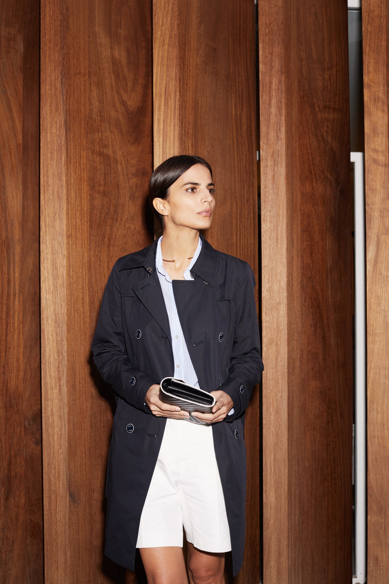 Woman in black coat and white shorts holding a black clutch against a wooden panel wall.
