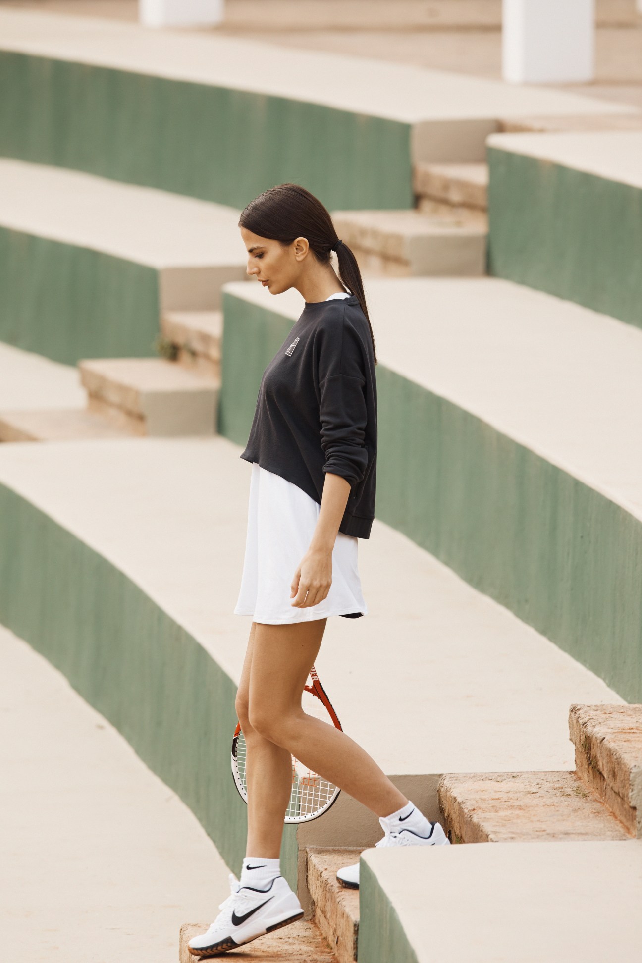 Model wearing Nike sweatshirt and dress with Nike trainers and socks, holding a Wilson racket on geometric steps.