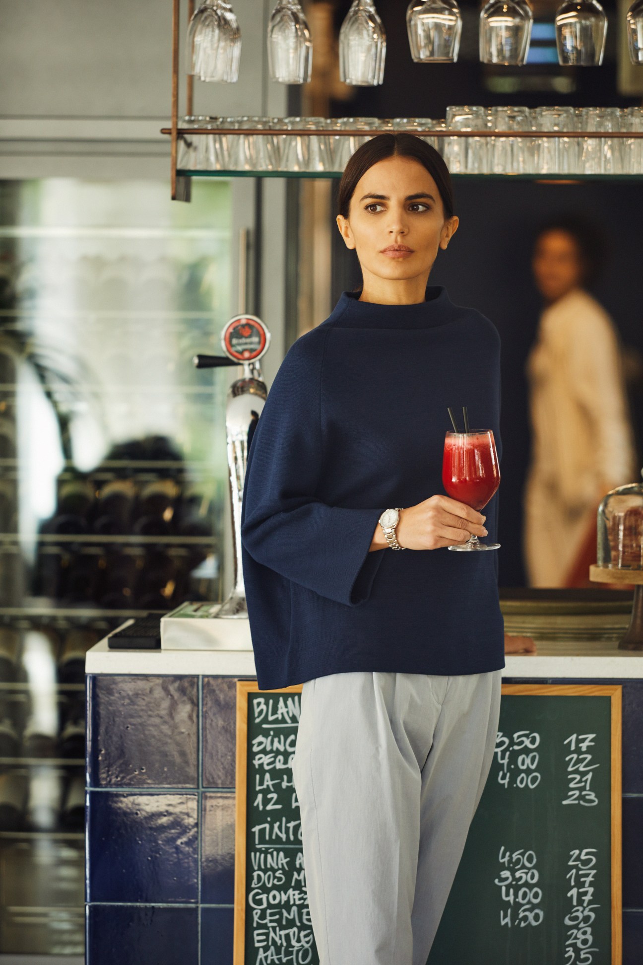 Woman in navy highneck jumper and light trousers holding a red drink at a bar.