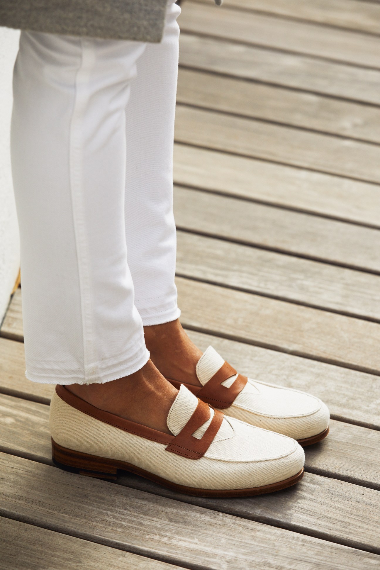 Person wearing white trousers and two-tone loafers with cream and brown leather on wooden decking.
