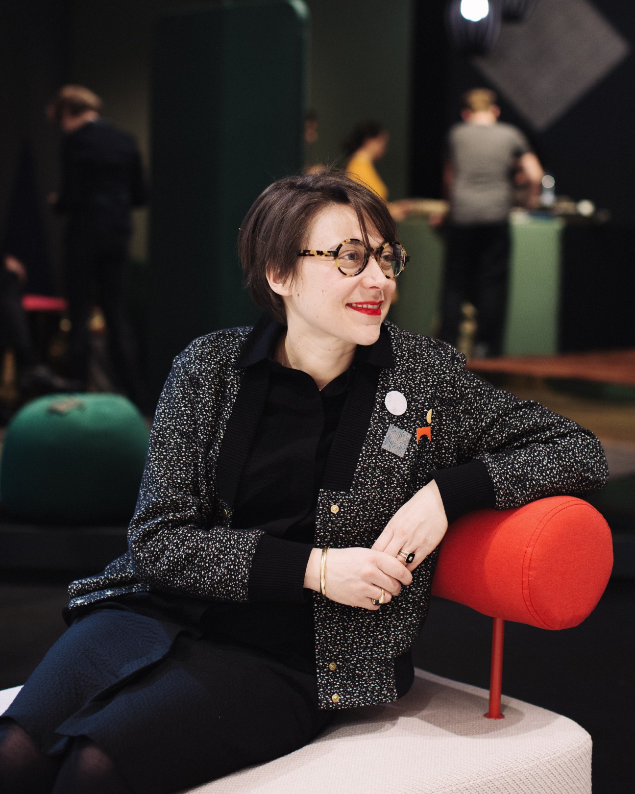 Woman with glasses wearing a sparkly blazer sitting on a red stool at IMM Cologne furniture fair