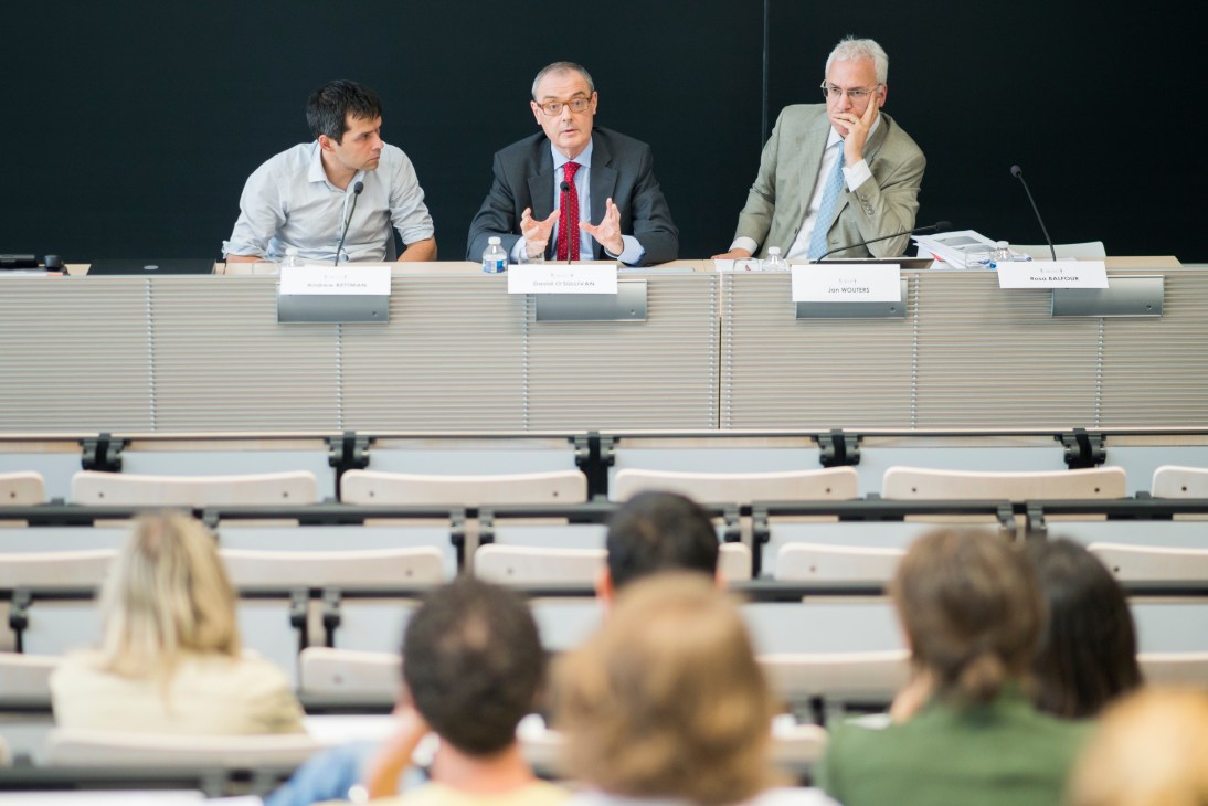 Panel of three men speaking at a lecture table in front of an audience in a large auditorium.