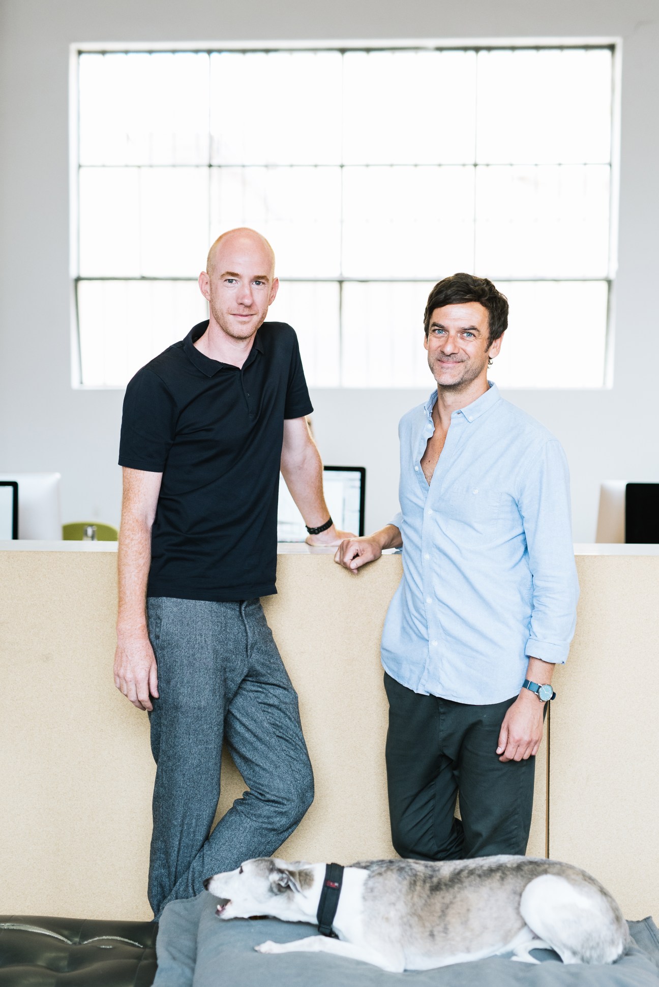 Two men stand behind a beige sofa with a whippet dog resting on the floor in front of them, in a bright studio with large windows.