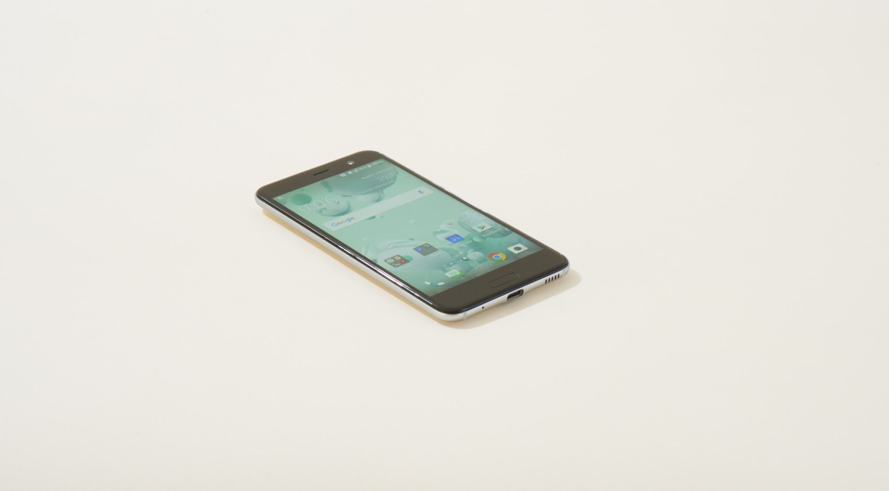 HTC U Play smartphone with pearlescent glass back in white with turquoise display screen shown at an angle against a light background.