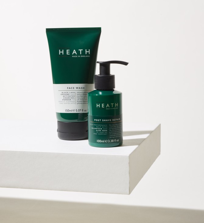 Heath face wash and post-shave repair skincare products in green bottles displayed on white geometric platform.