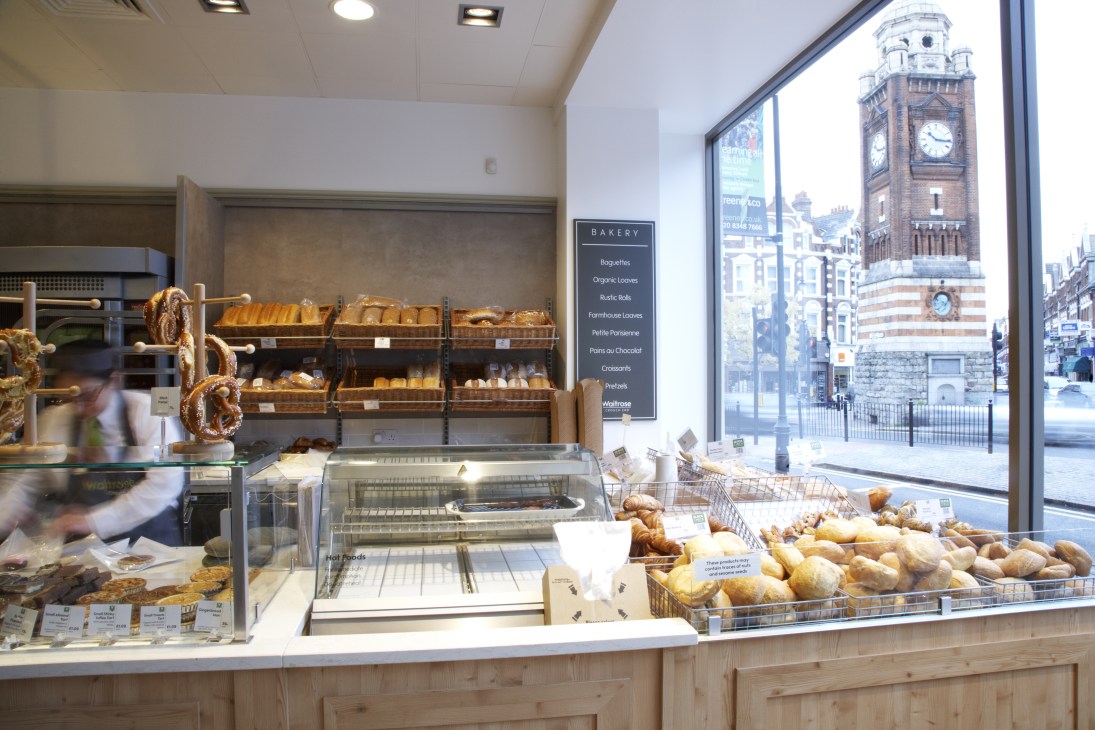 Waitrose bakery counter displays fresh bread, pastries and baked goods with a clock tower visible through the window.