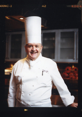 Chef in white uniform and tall white chef's hat smiling at camera in restaurant kitchen.