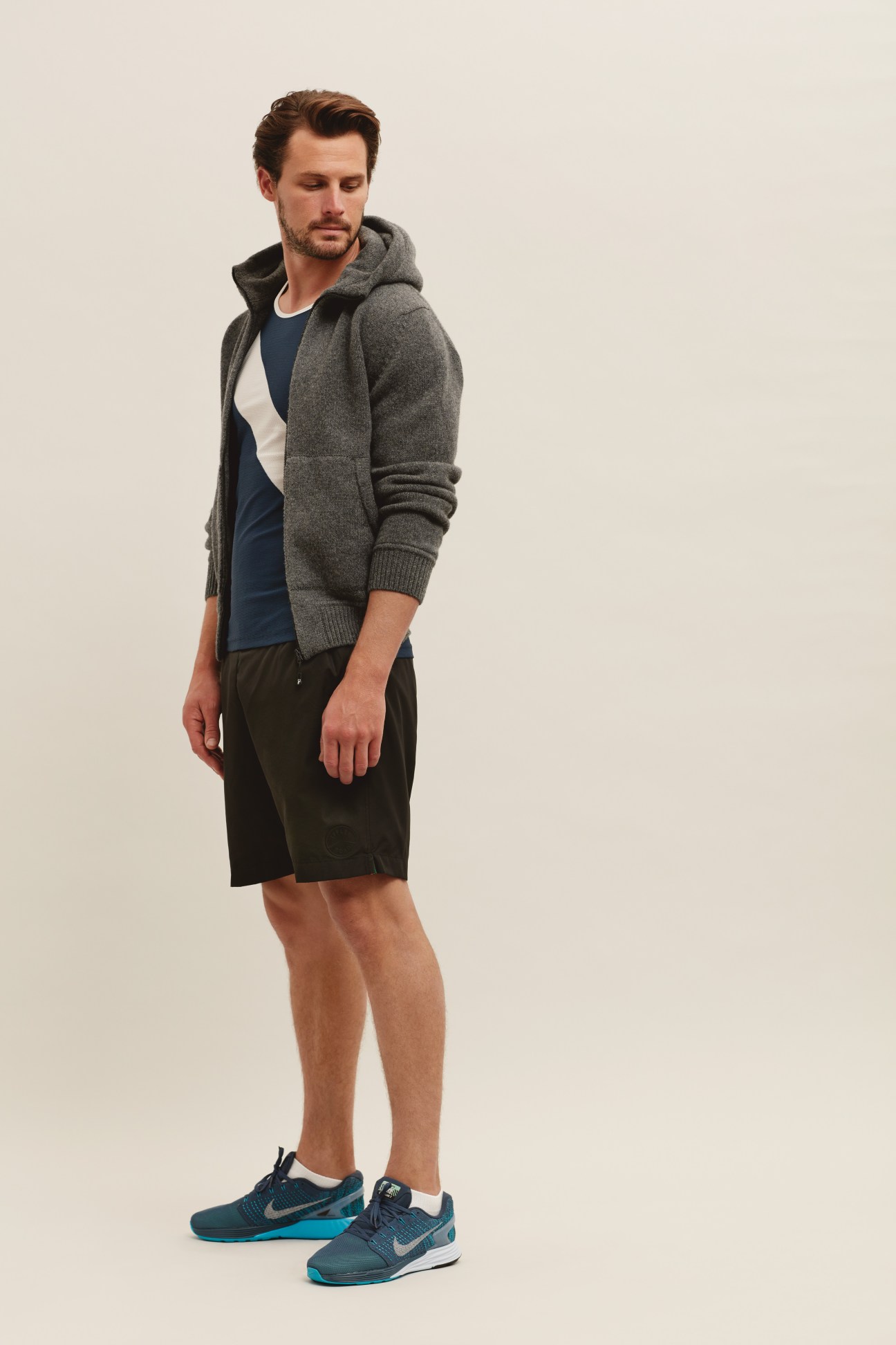 Man wearing grey zip-up hoodie, navy t-shirt, black shorts, and blue trainers standing against neutral background.