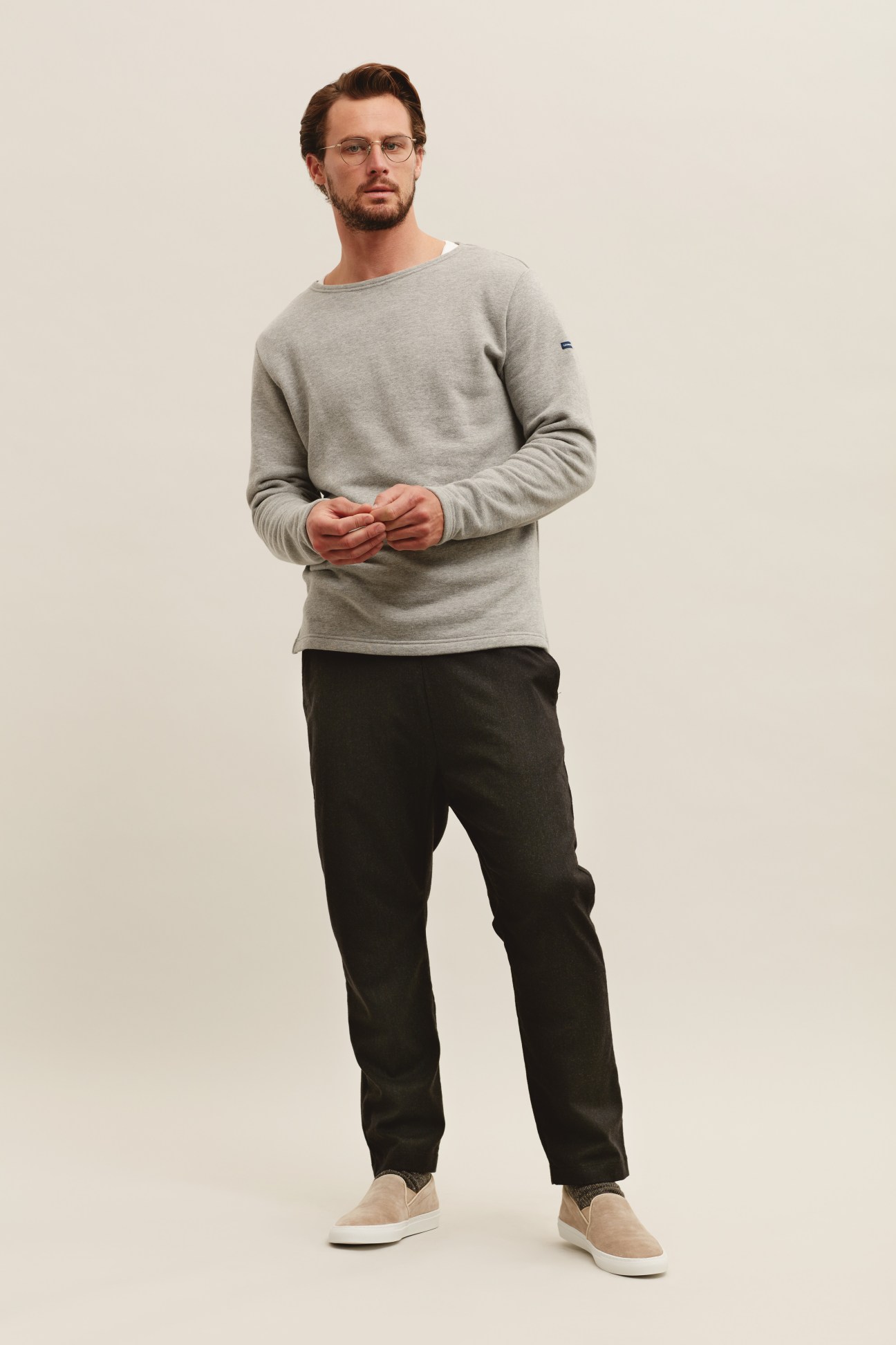 Man wearing grey sweatshirt, black trousers, and beige slip-on shoes against neutral background.
