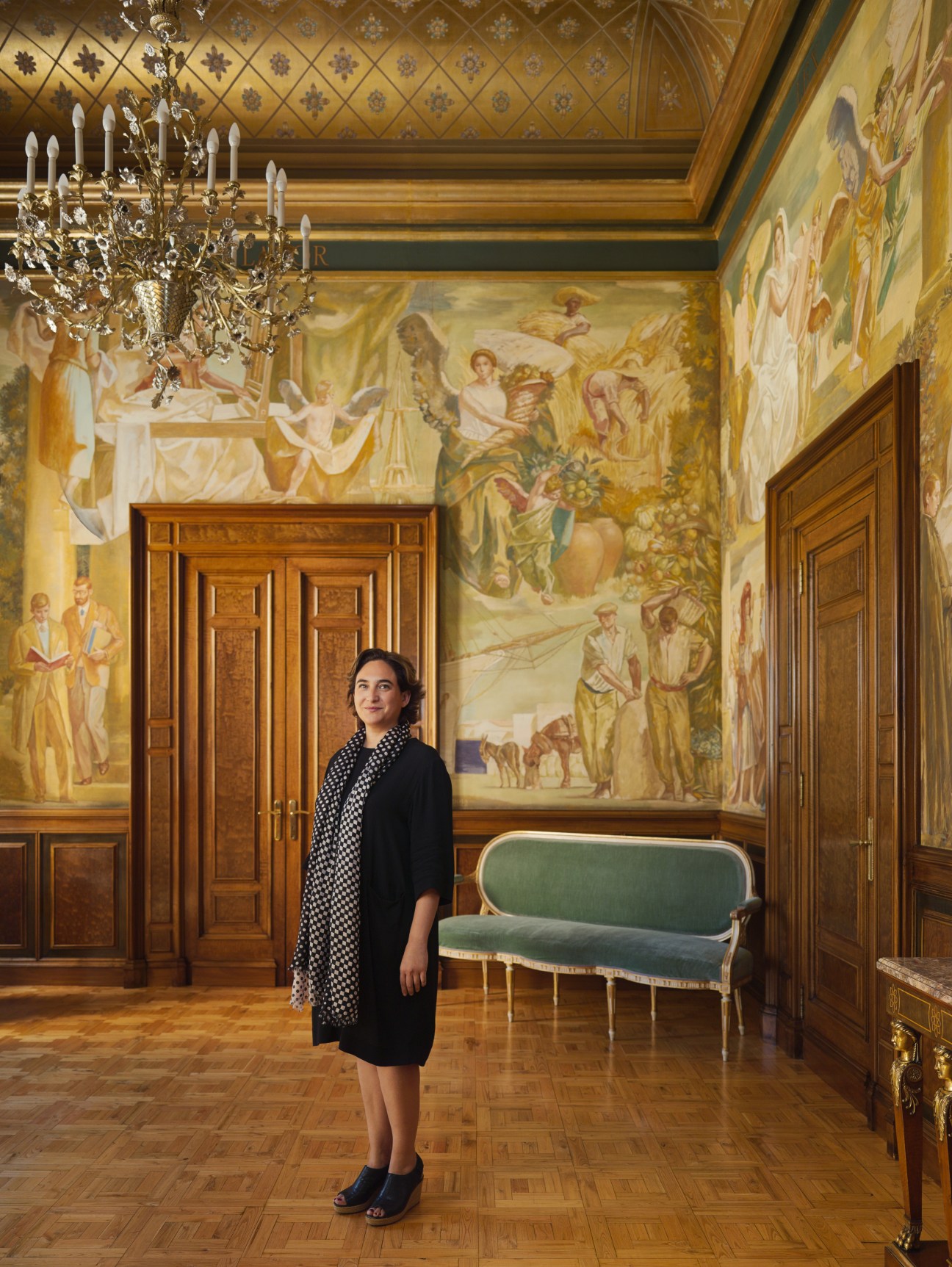 Ada Colau stands in an ornate room with frescoed walls, classical chandelier, and period furniture.