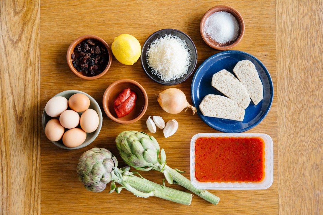 Ingredients for carciofi tappati arranged on a wooden surface including artichokes, eggs, bread, parmesan, sultanas and tomato passata.