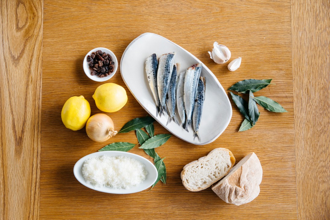 Ingredients for Sarde a beccafico laid out on a wooden surface: sardine fillets, lemons, onion, garlic, sultanas, breadcrumbs, and bay leaves.