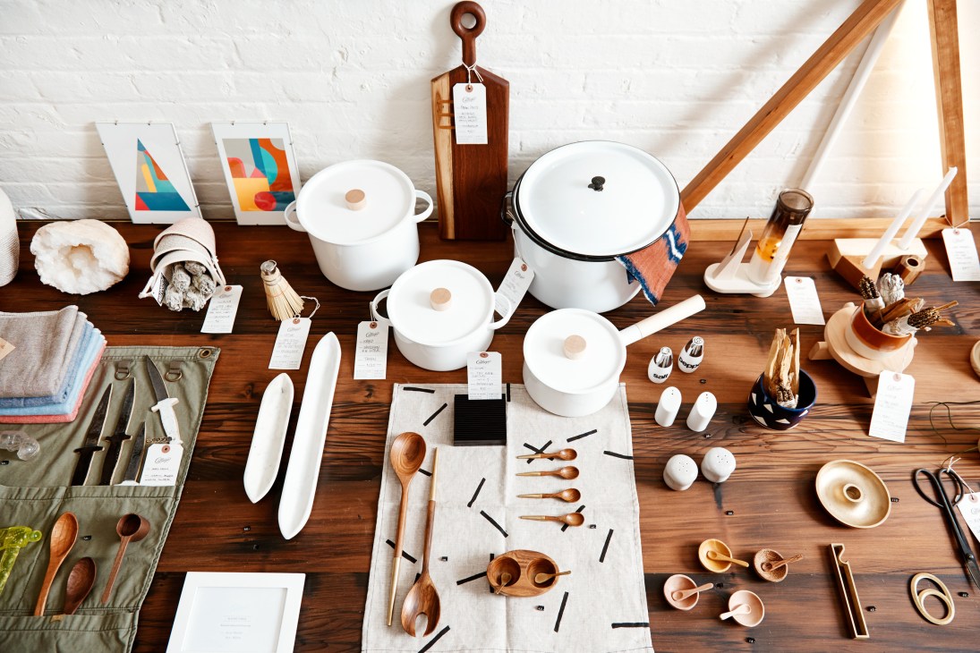 Overhead view of curated homeware display featuring white cookware, wooden utensils, ceramics, and textiles on wooden table.
