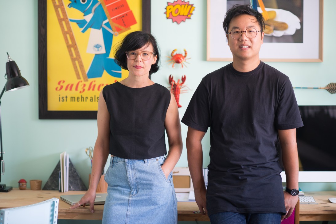 Daniel To and Emma Aiston stand in their Adelaide studio surrounded by colorful graphic designs and artwork on the walls.