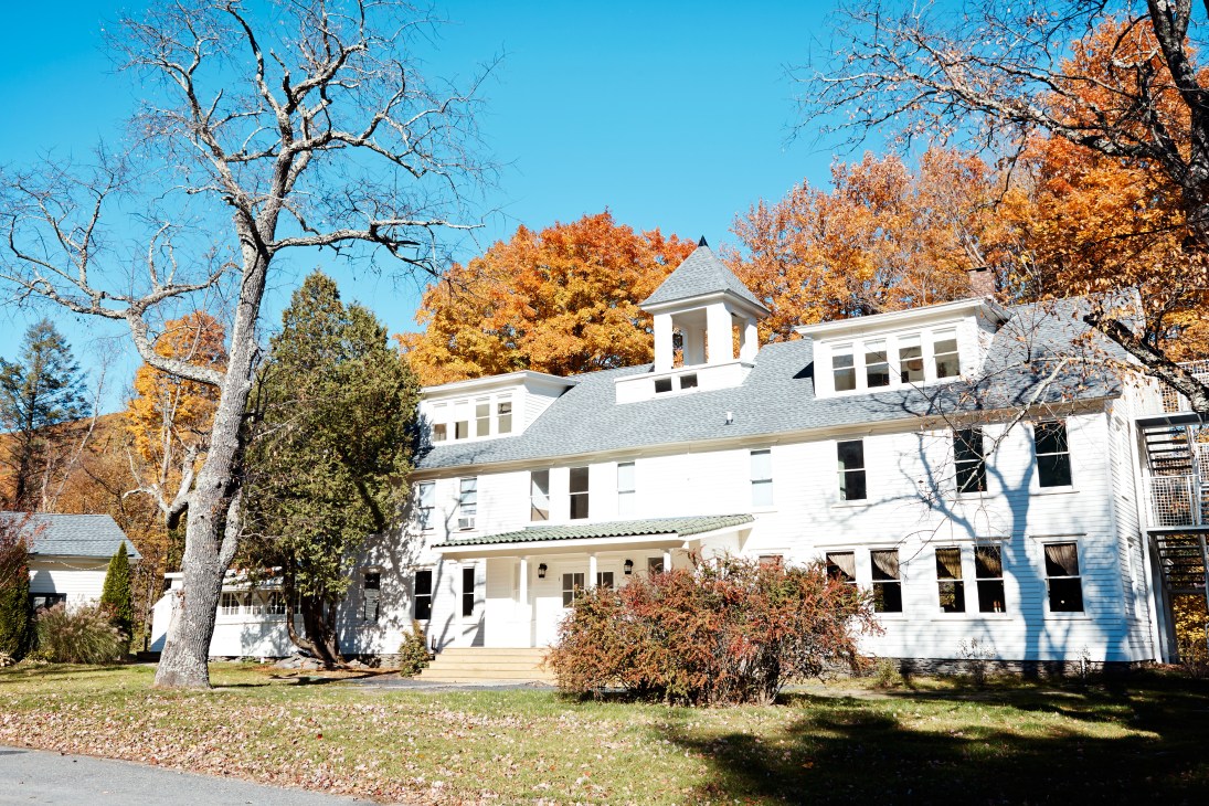 White wooden inn with gray roof and cupolas surrounded by autumn foliage and bare trees.