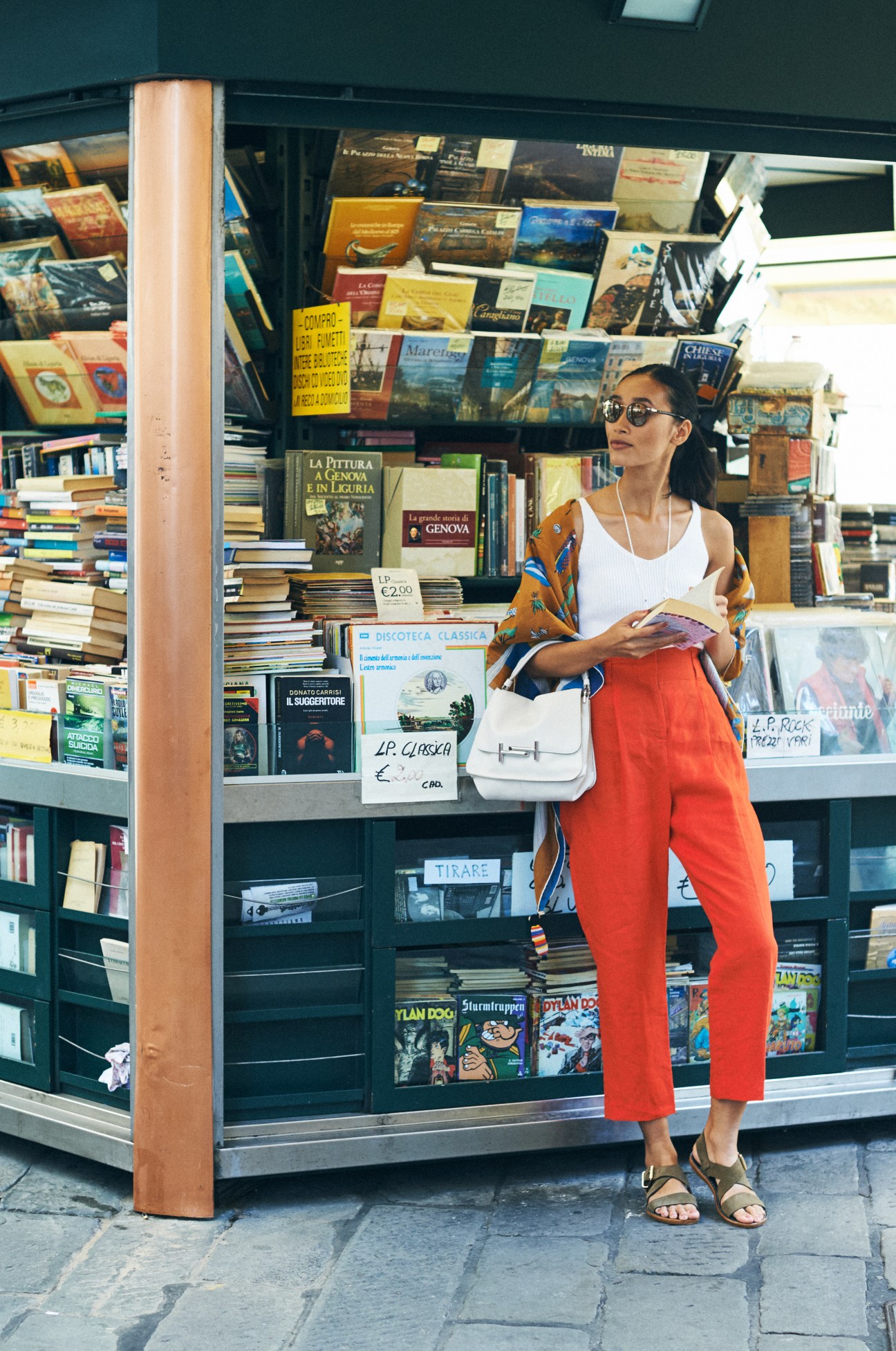 Woman in white vest, red trousers and sandals browsing books at a Genoa bookshop window.